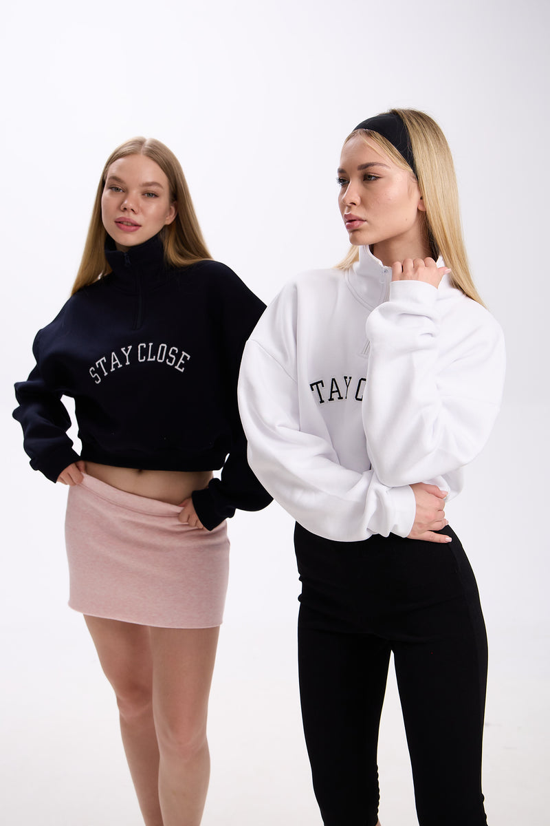 Two women wearing cropped half-zip hoodies with 'STAY CLOSE' text – one in navy paired with a pastel pink mini skirt, the other in white paired with black leggings, both posing on a plain background