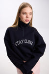 Navy cropped half-zip hoodie with white 'STAY CLOSE' embroidery – close-up detail shot styled with pastel pink mini skirt, minimal streetwear look on plain backdrop (dark navy shade similar to black)
