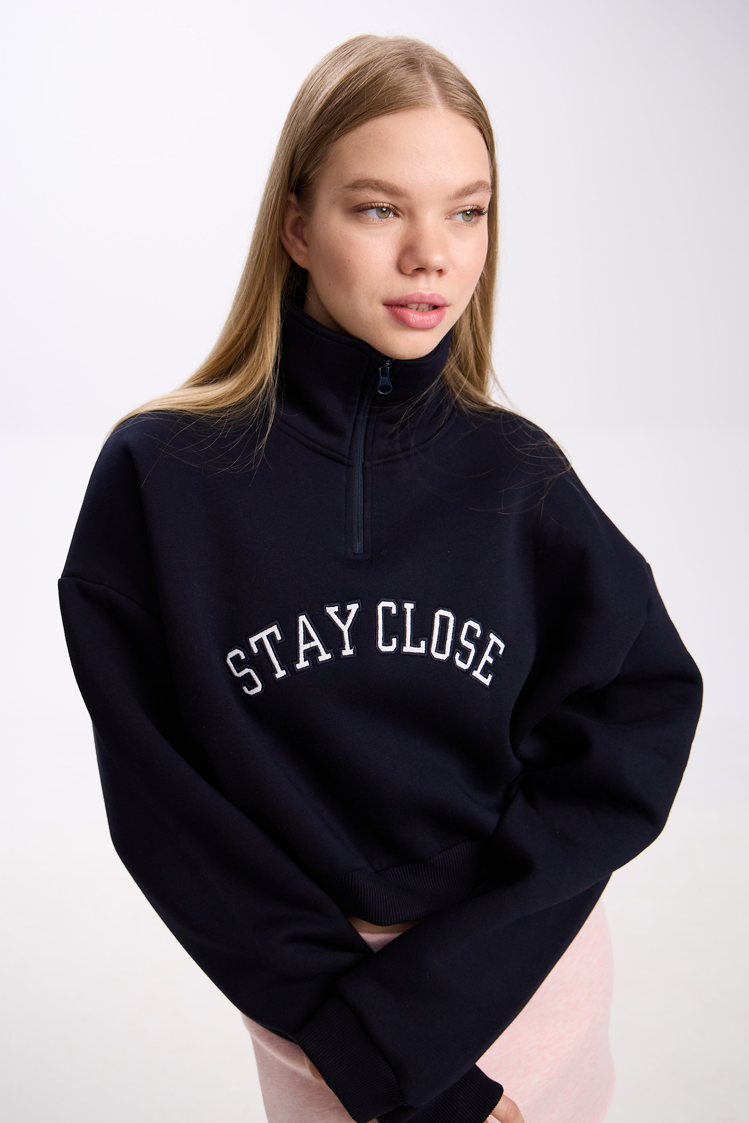 Navy cropped half-zip hoodie with white 'STAY CLOSE' embroidery – close-up detail shot styled with pastel pink mini skirt, minimal streetwear look on plain backdrop (dark navy shade similar to black)