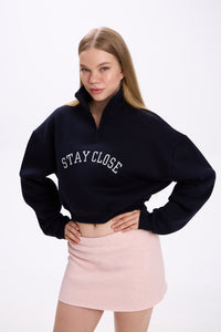 Navy cropped half-zip hoodie with bold white 'STAY CLOSE' embroidery, styled with pastel pink mini skirt – minimal streetwear outfit, hands-on-hips pose on plain backdrop (dark navy shade close to black)