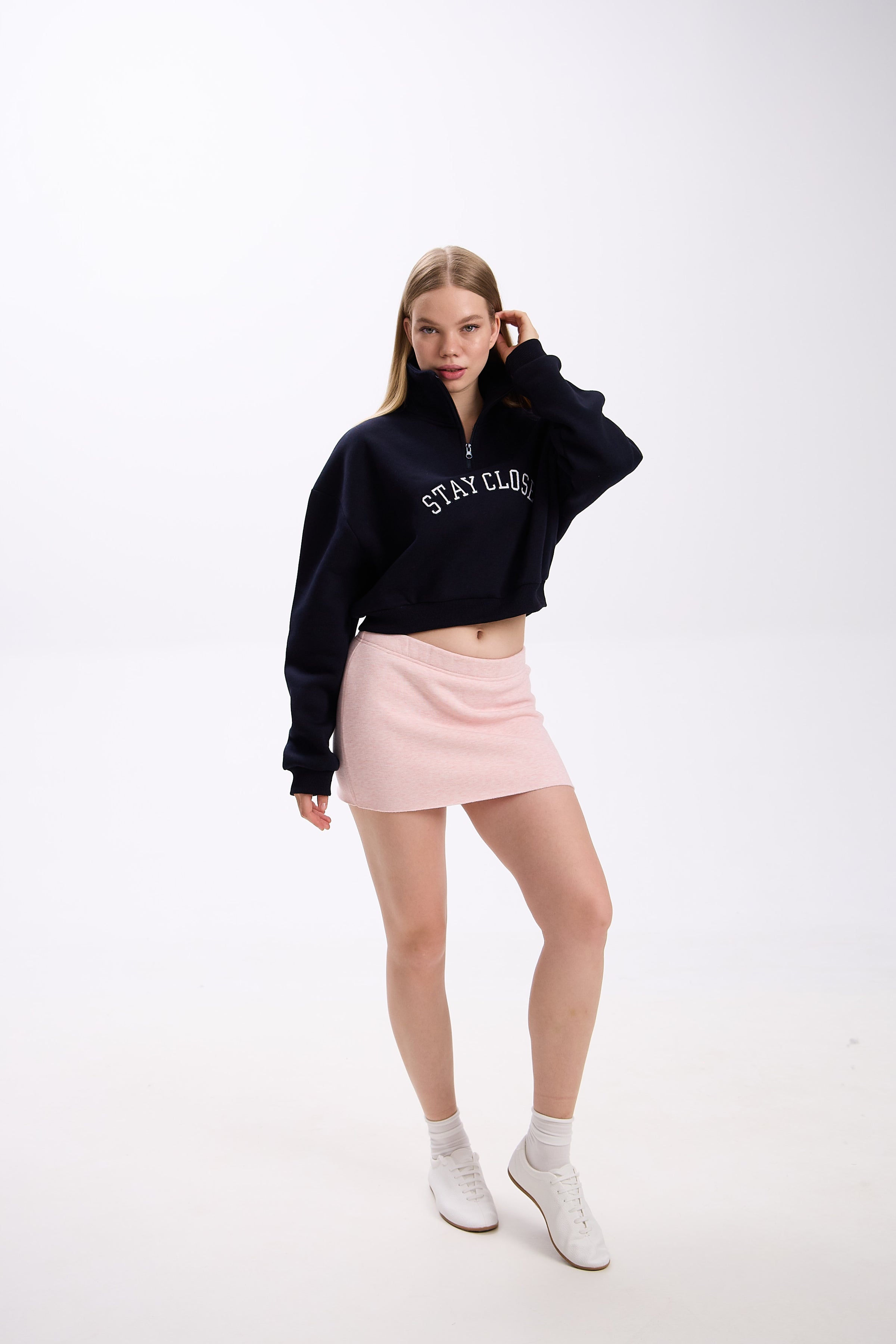 Navy cropped half-zip hoodie with bold white 'STAY CLOSE' embroidery, styled with pastel pink mini skirt and casual white sneakers – full-body streetwear look, hair-touch pose on plain backdrop (dark navy shade close to black)