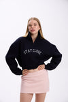 Navy cropped half-zip hoodie with white 'STAY CLOSE' embroidery, styled with pastel pink mini skirt – minimal streetwear outfit, confident pose with hand on waist on plain backdrop (dark navy shade close to black)