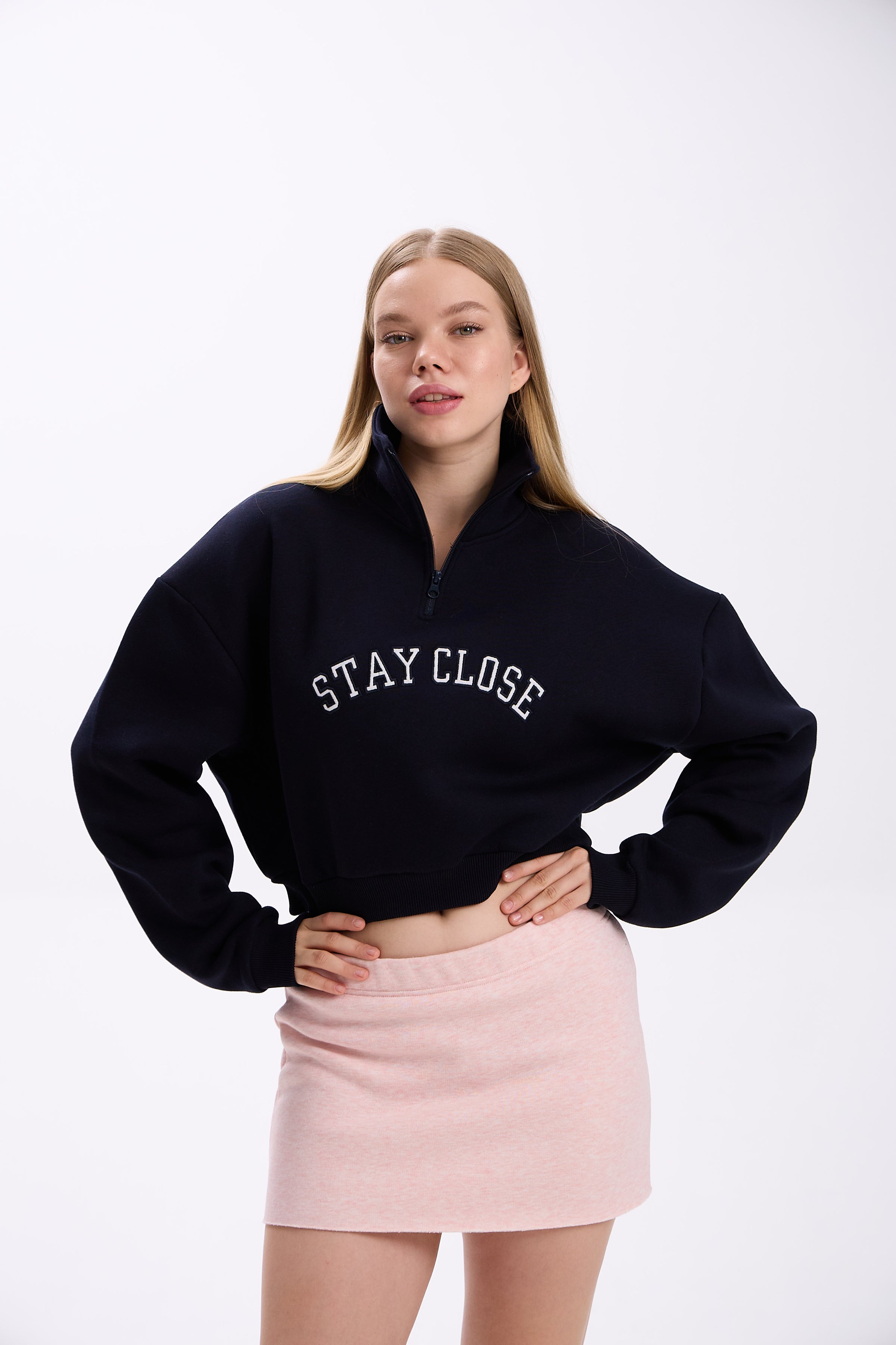Navy cropped half-zip hoodie with white 'STAY CLOSE' embroidery, styled with pastel pink mini skirt – minimal streetwear outfit, confident pose with hand on waist on plain backdrop (dark navy shade close to black)