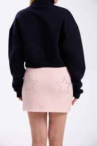 Back view of navy cropped half-zip hoodie styled with pastel pink mini skirt featuring silver star stud details – minimal streetwear outfit on plain backdrop (dark navy shade close to black)