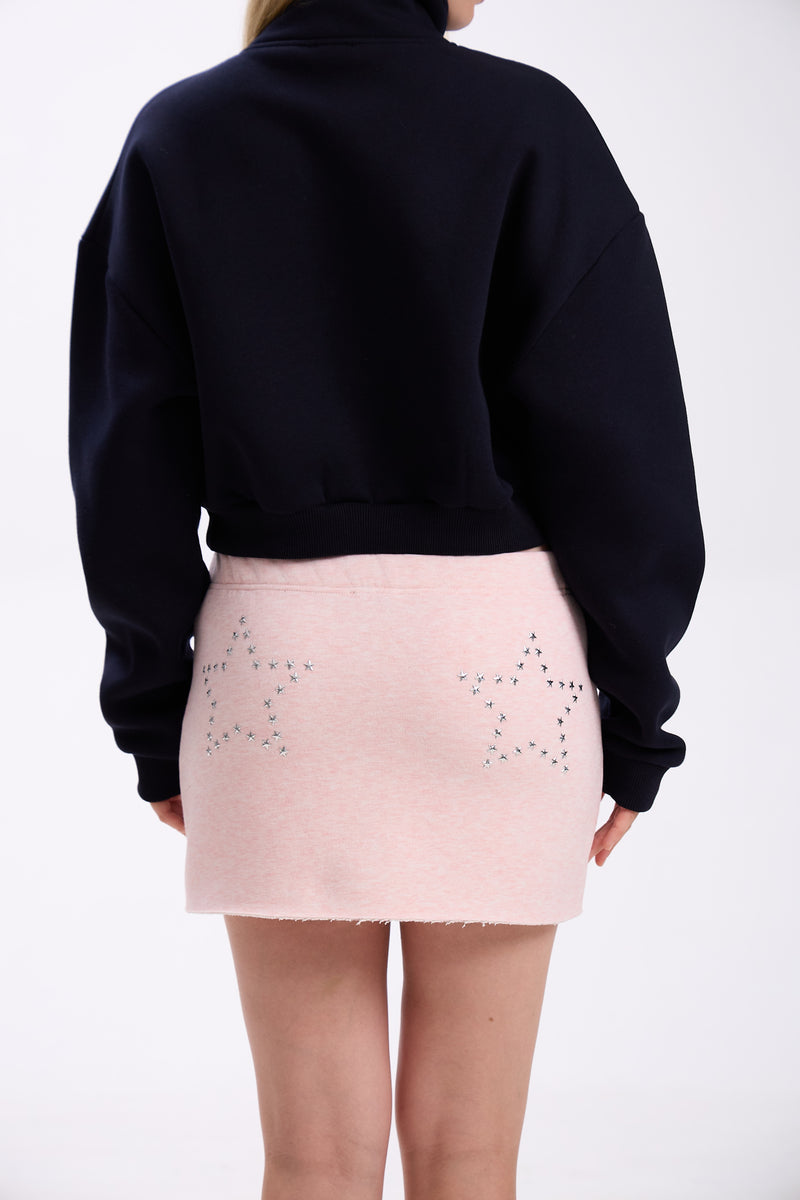 Back view of navy cropped half-zip hoodie styled with pastel pink mini skirt featuring silver star stud details – minimal streetwear outfit on plain backdrop (dark navy shade close to black)