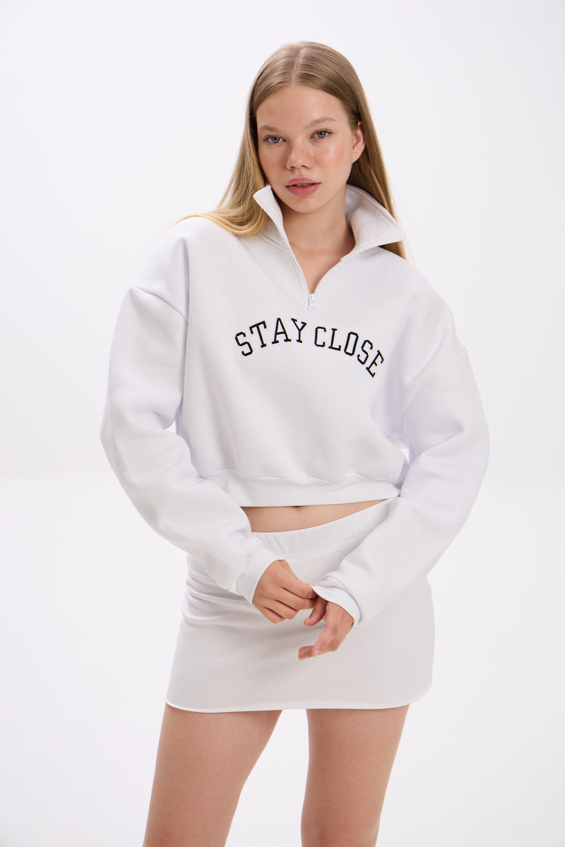 White cropped half-zip hoodie with 'STAY CLOSE' chest print, styled with matching white mini skirt – minimal streetwear look on plain backdrop