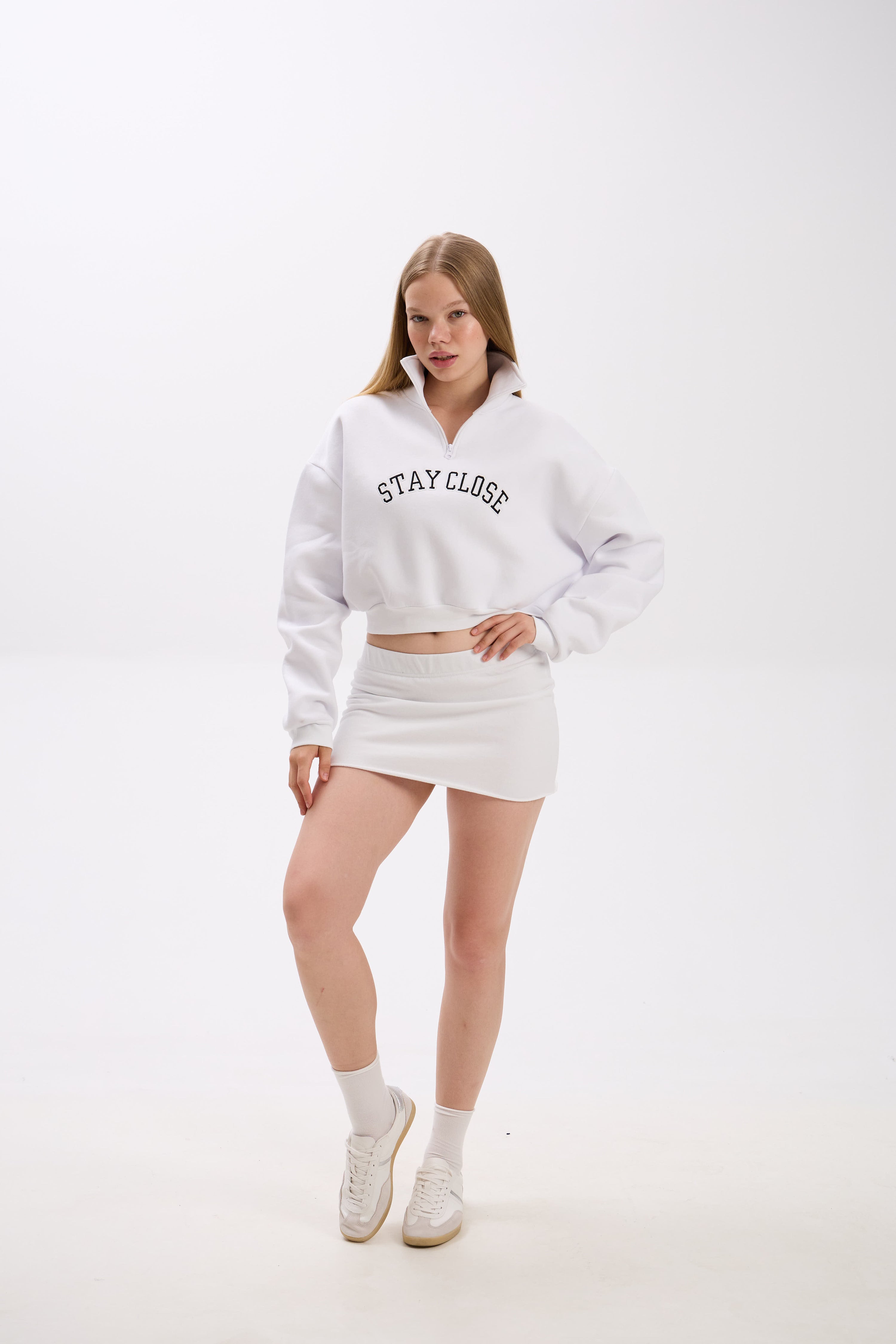 Person wearing a white sweatshirt with 'STAY CLOSE' chest embroidery, paired with white mini skirt, socks, and sneakers.