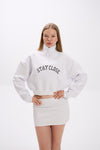 White cropped half-zip hoodie with 'STAY CLOSE' chest embroidery, styled with matching white mini skirt – minimal streetwear outfit, hands-on-hips pose on plain backdrop