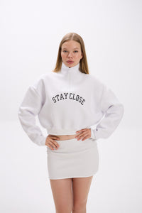 White cropped half-zip hoodie with 'STAY CLOSE' chest embroidery, styled with matching white mini skirt – minimal streetwear outfit, hands-on-hips pose on plain backdrop