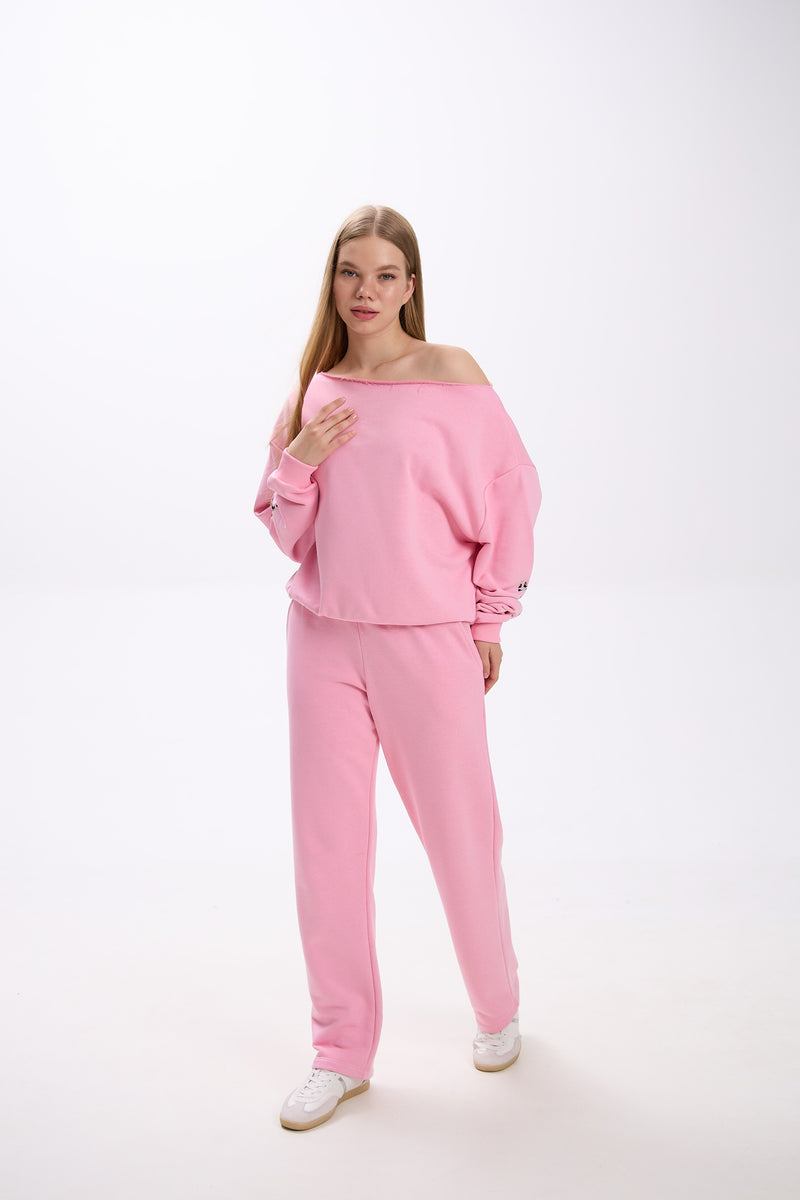 Model wearing a pink loungewear set with an off-shoulder oversized sweatshirt and straight-leg sweatpants, styled with white sneakers.