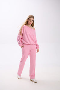 Model in a pink Stay Close loungewear set featuring an off-shoulder oversized sweatshirt with logo detail on the sleeve and matching straight-leg sweatpants, styled with white sneakers.