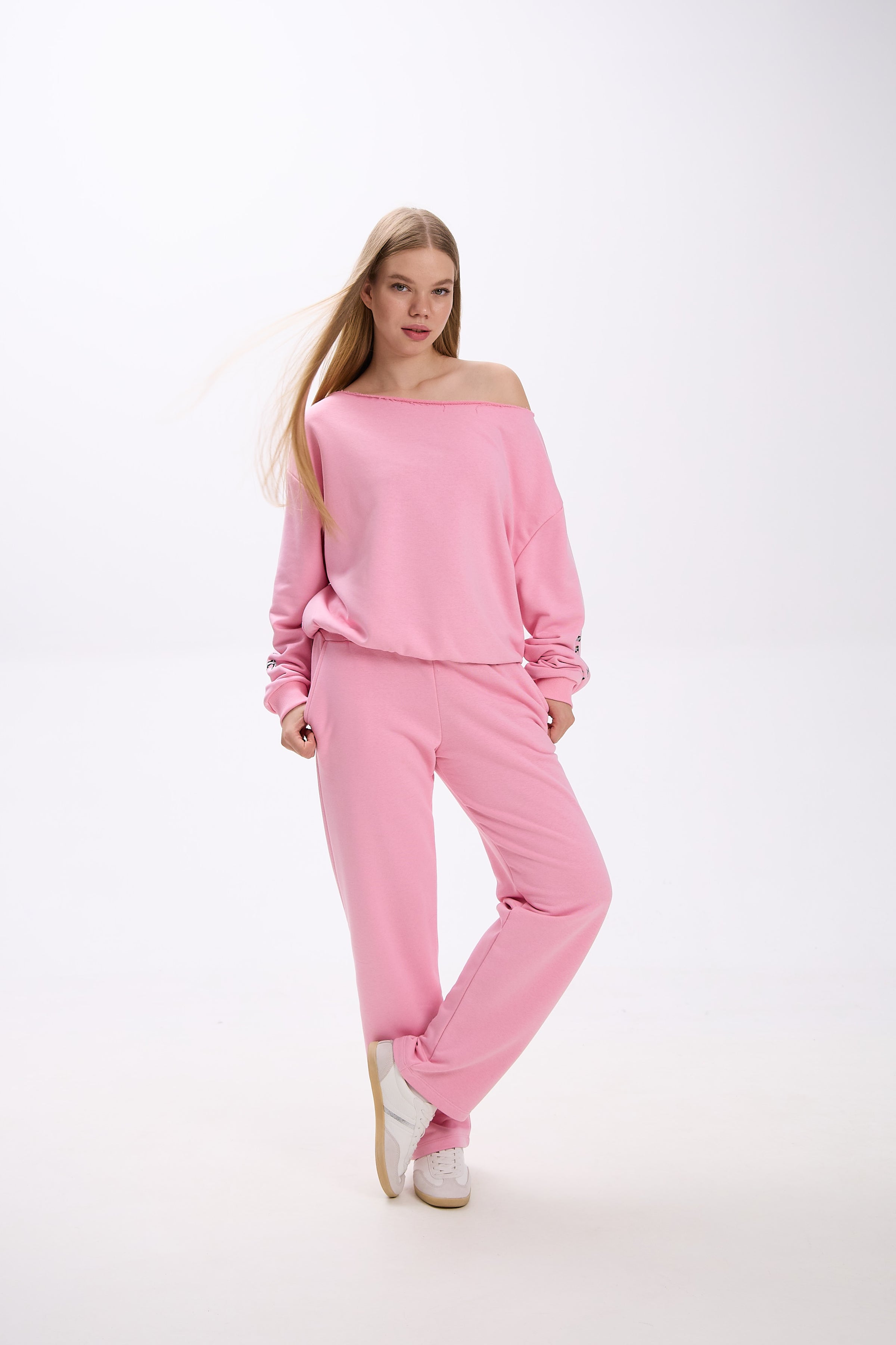 Model in a pink Stay Close loungewear set featuring an off-shoulder oversized sweatshirt with logo detail on the sleeve and matching straight-leg sweatpants, styled with white sneakers.