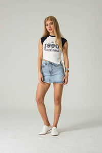 Woman wearing a black and white baseball shirt with '1990 England' text, light blue denim skirt, and white sneakers on a plain background.