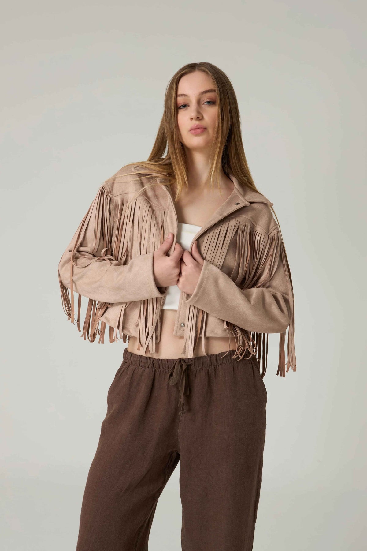Woman wearing a beige fringe jacket and brown pants against a plain background.