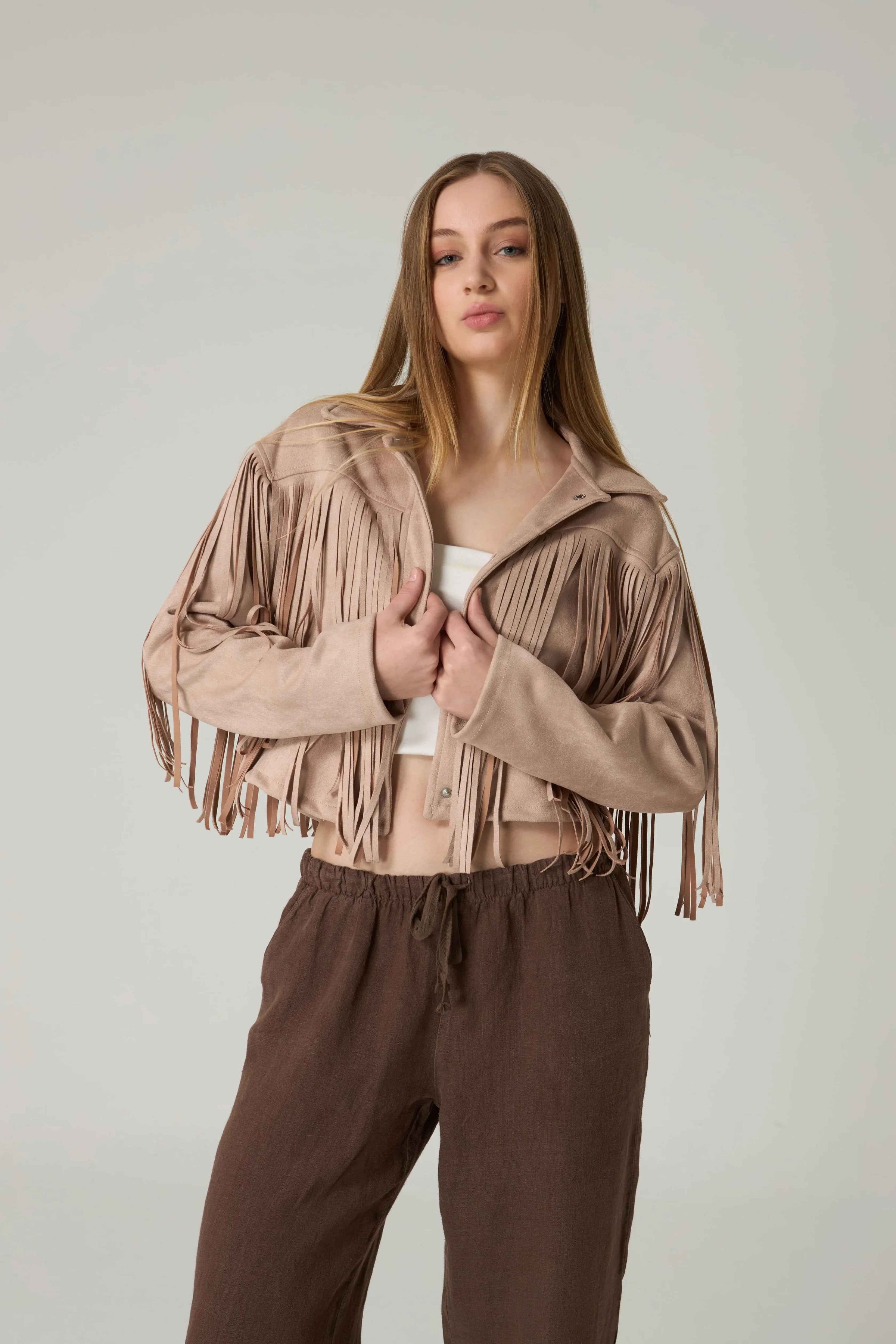 Woman wearing a beige fringe jacket and brown pants against a plain background.