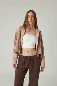 Woman wearing a beige jacket, white top, and brown pants on a plain background.