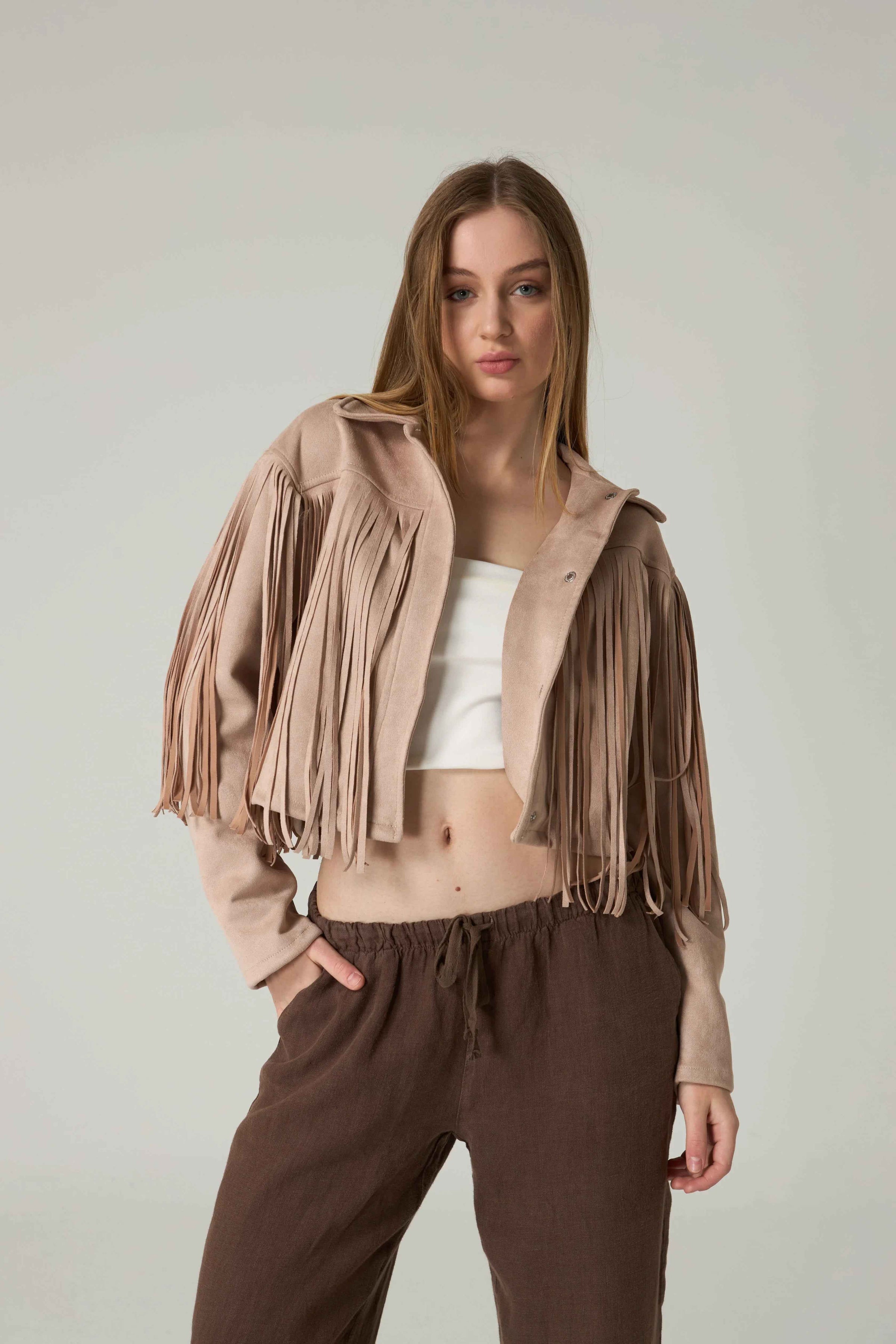 Woman wearing a beige fringe jacket and brown pants against a plain background.