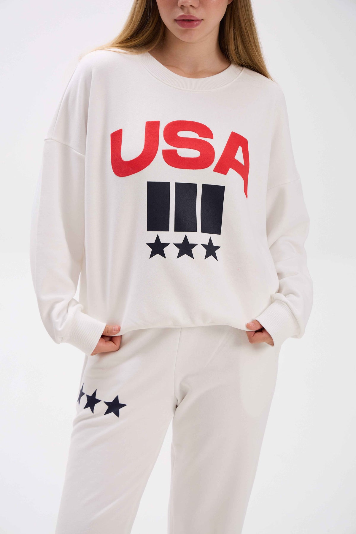 Person wearing a white sweatshirt with 'USA' and star graphics on a plain background.