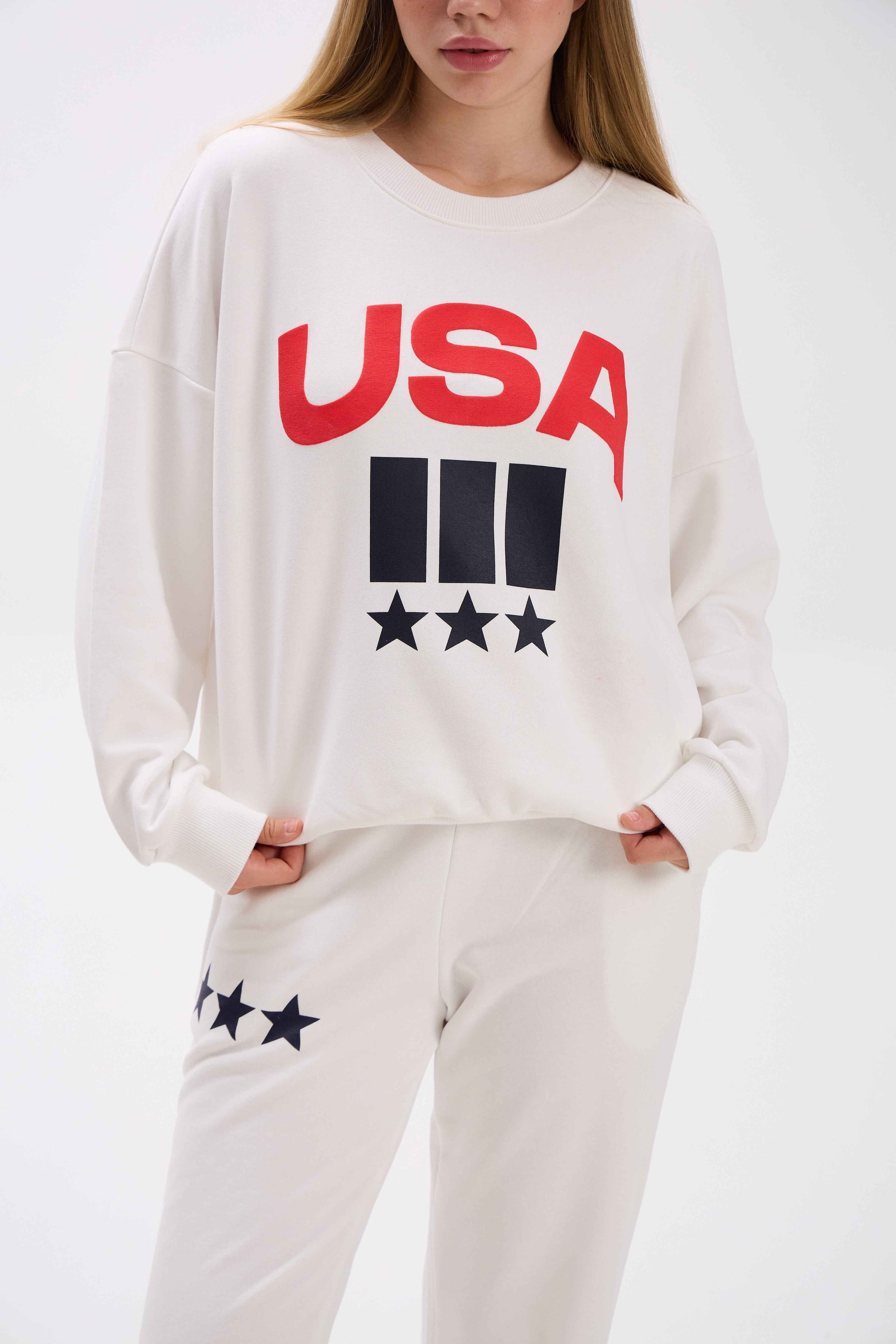 Person wearing a white sweatshirt with 'USA' and star graphics on a plain background.