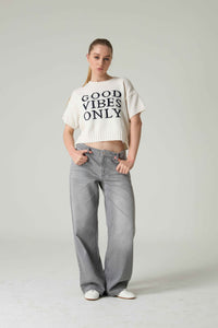 Person wearing a white 'Good Vibes Only' shirt and gray jeans on a plain background.
