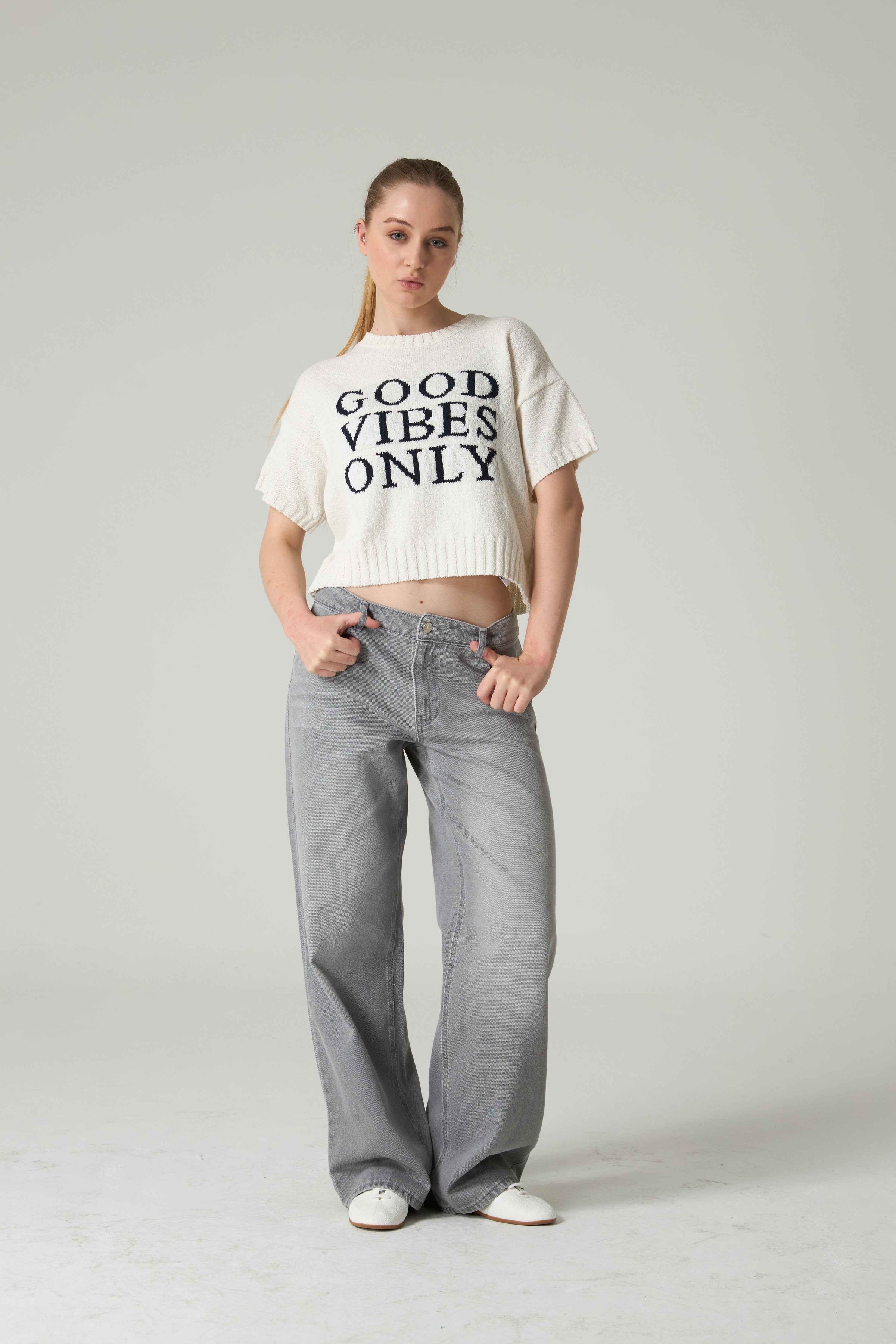 Person wearing a white 'Good Vibes Only' shirt and gray jeans on a plain background.