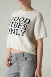 Person wearing a white knit sweater with 'GOOD VIBES ONLY' text on a plain background.