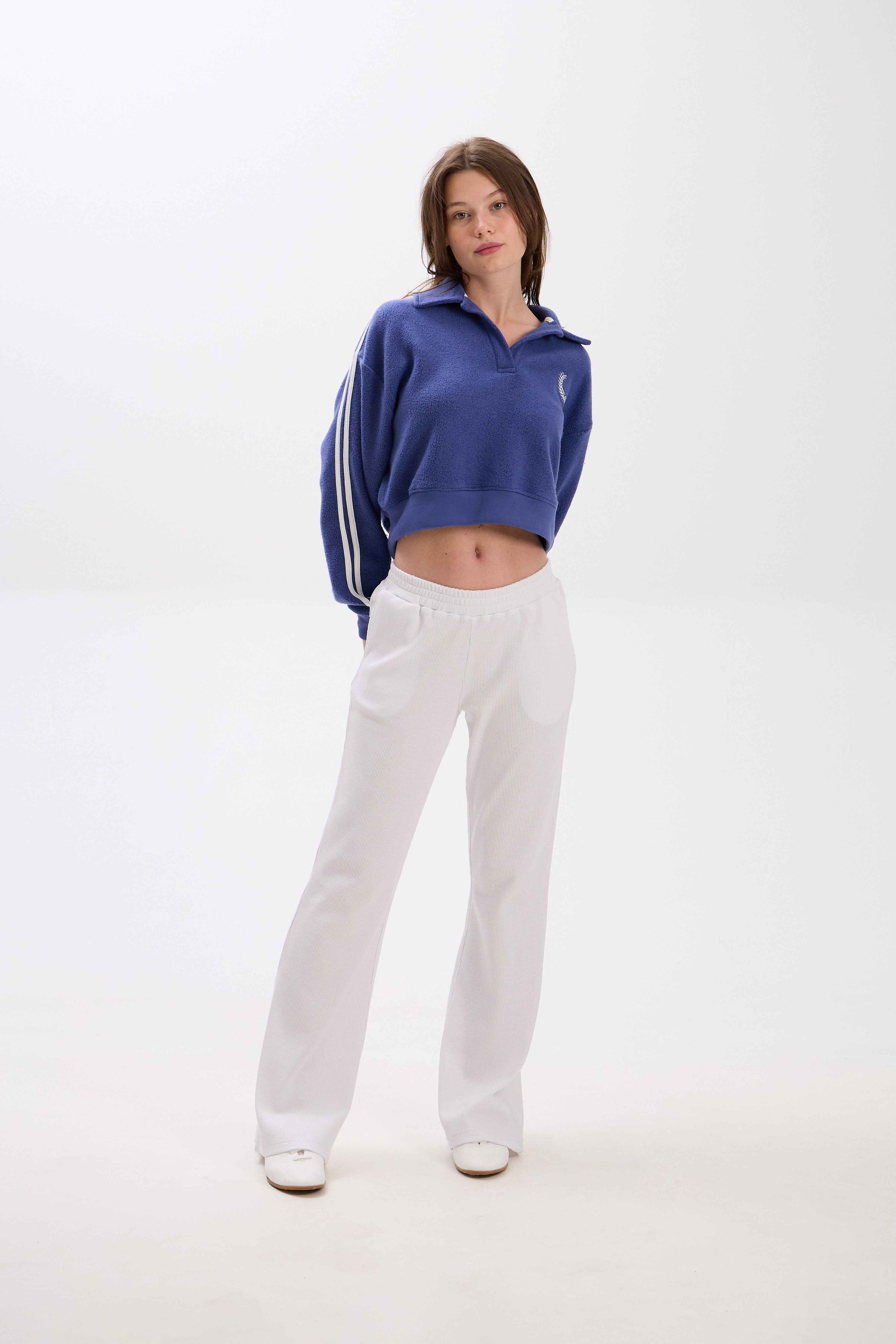 Person wearing a blue cropped sweatshirt with white stripes and white pants on a white background.