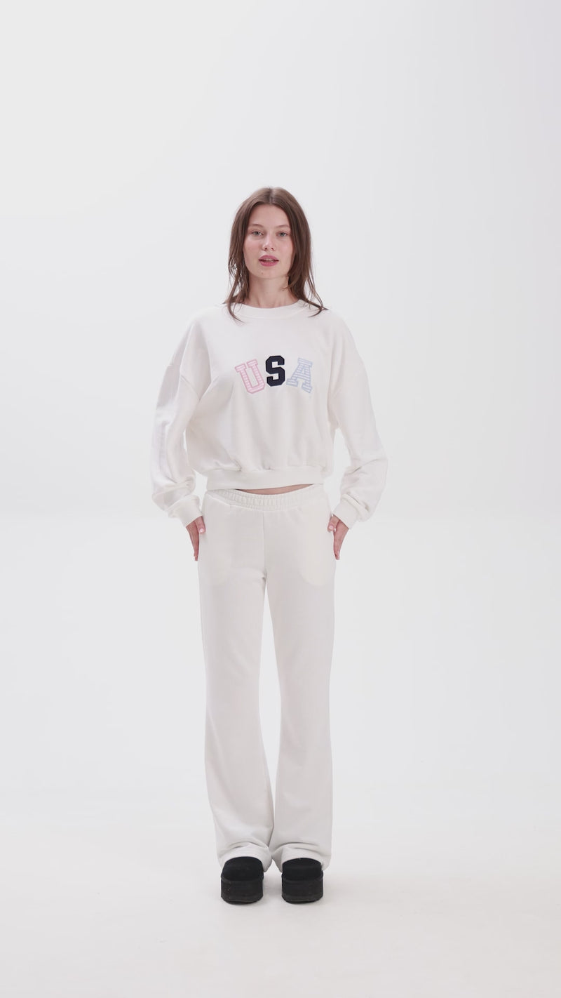 White sweatpants with elastic waistband, side pockets, and multicolor USA lettering on the back, shown in motion for a casual look.