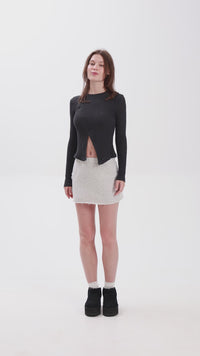 Person wearing a fitted black long-sleeve top with a front slit, paired with a light grey mini skirt, white socks, and black shoes.