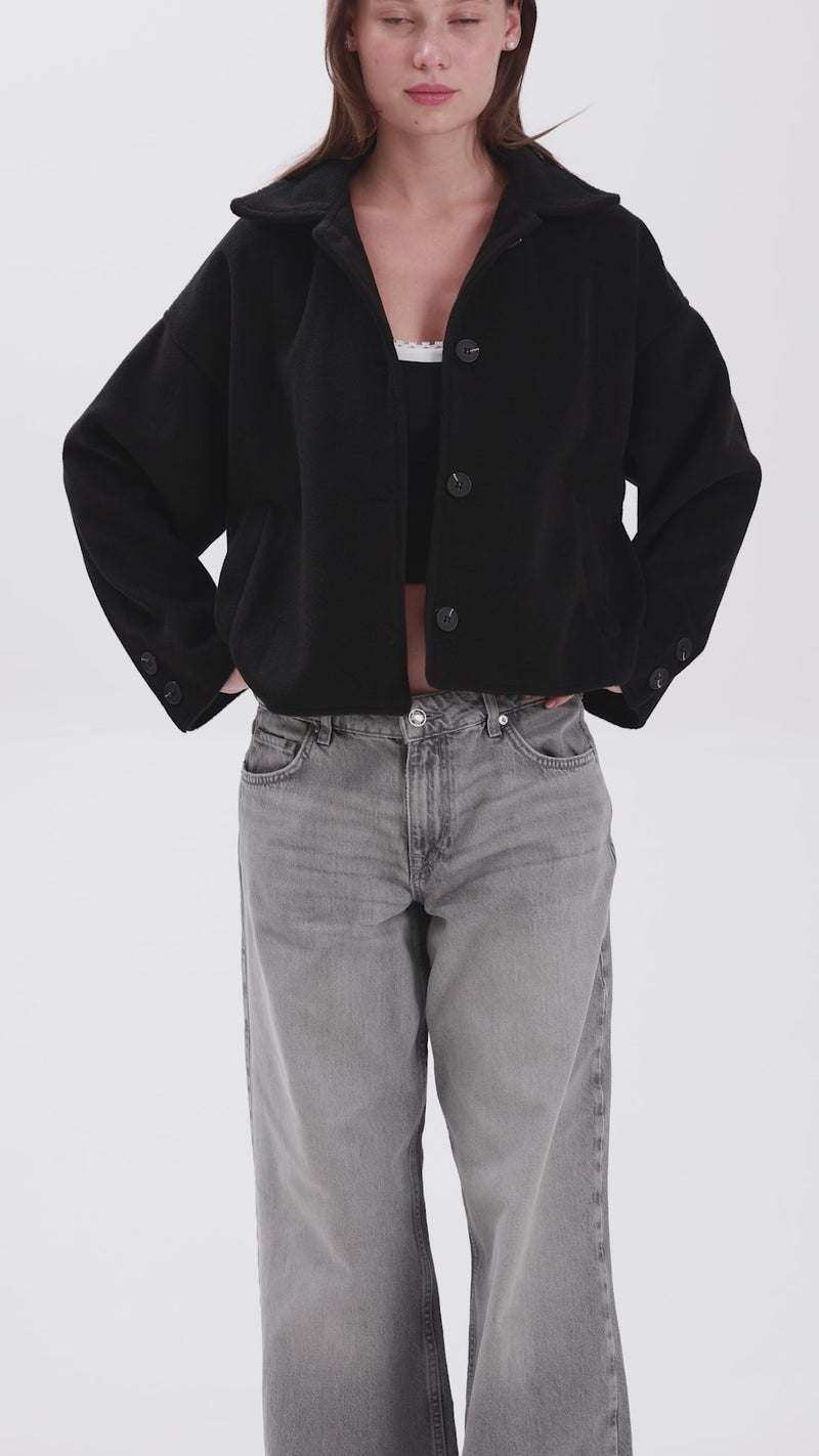 Woman wearing a cropped black button-up jacket with wide sleeves, styled with high-waisted loose-fit grey jeans, standing against a white background.