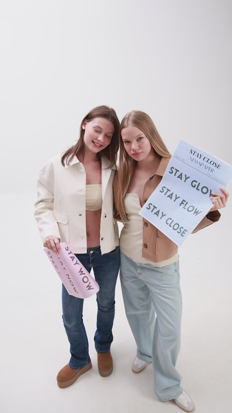 Two young women standing together in casual outfits, holding large paper sheets with “STAY CLOSE,” “STAY FLOW,” and “STAY NOW” printed on them.