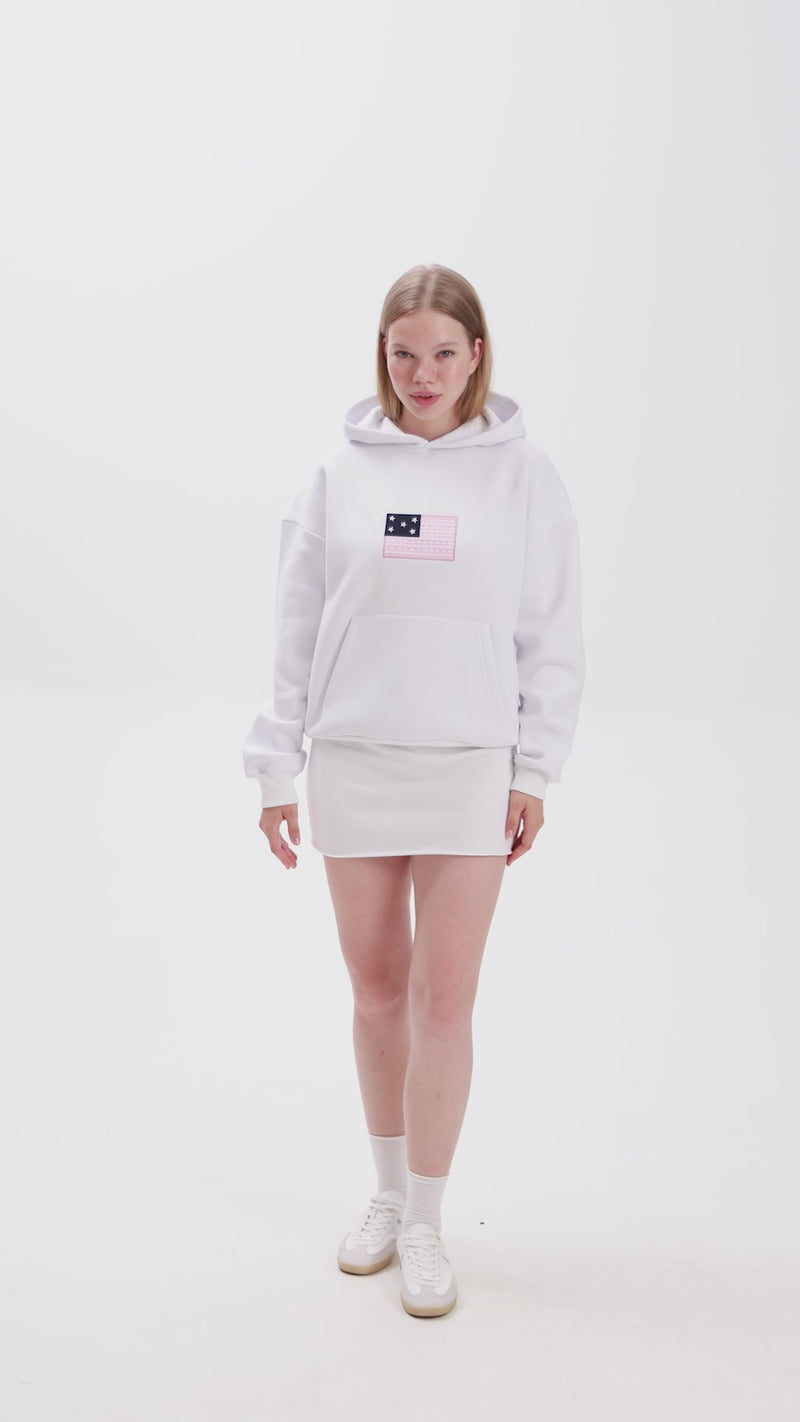 Video of a woman wearing a white hoodie with a pink and navy star patch design on the chest, paired with a matching white mini skirt and white sneakers. She poses by placing her hands in the hoodie pocket and pulling up the hood, showcasing the outfit details against a plain background.