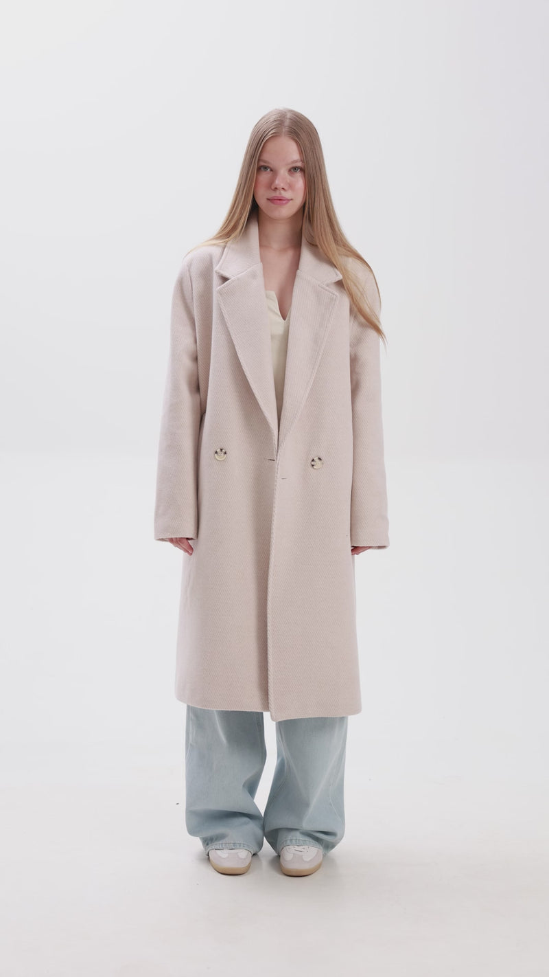 Woman wearing a long beige overcoat with large lapels, paired with light blue wide-leg jeans and white sneakers, standing against a white background.