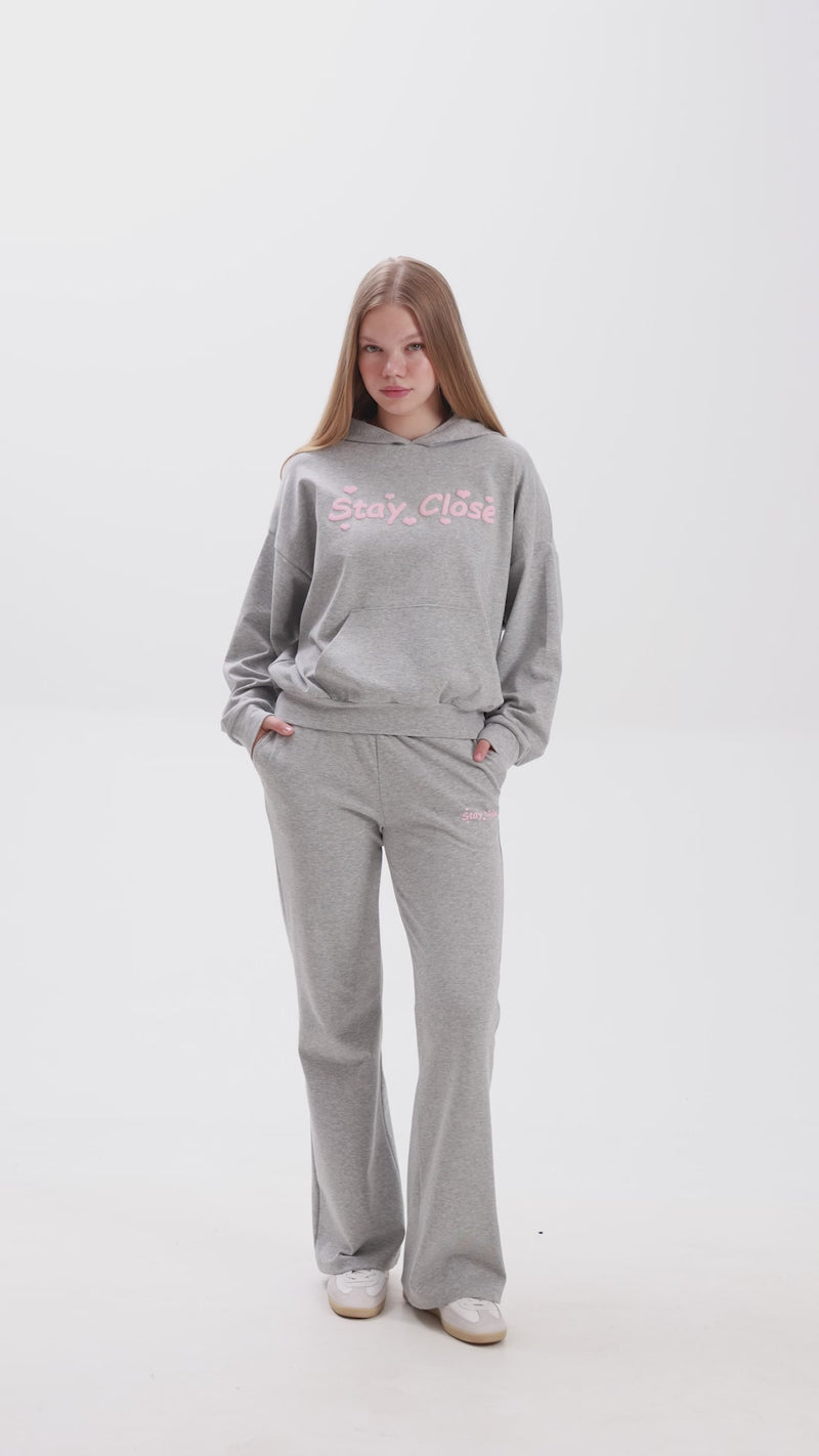 Person wearing grey melange sweatpants and matching hoodie with pink “Stay Close” embroidery.