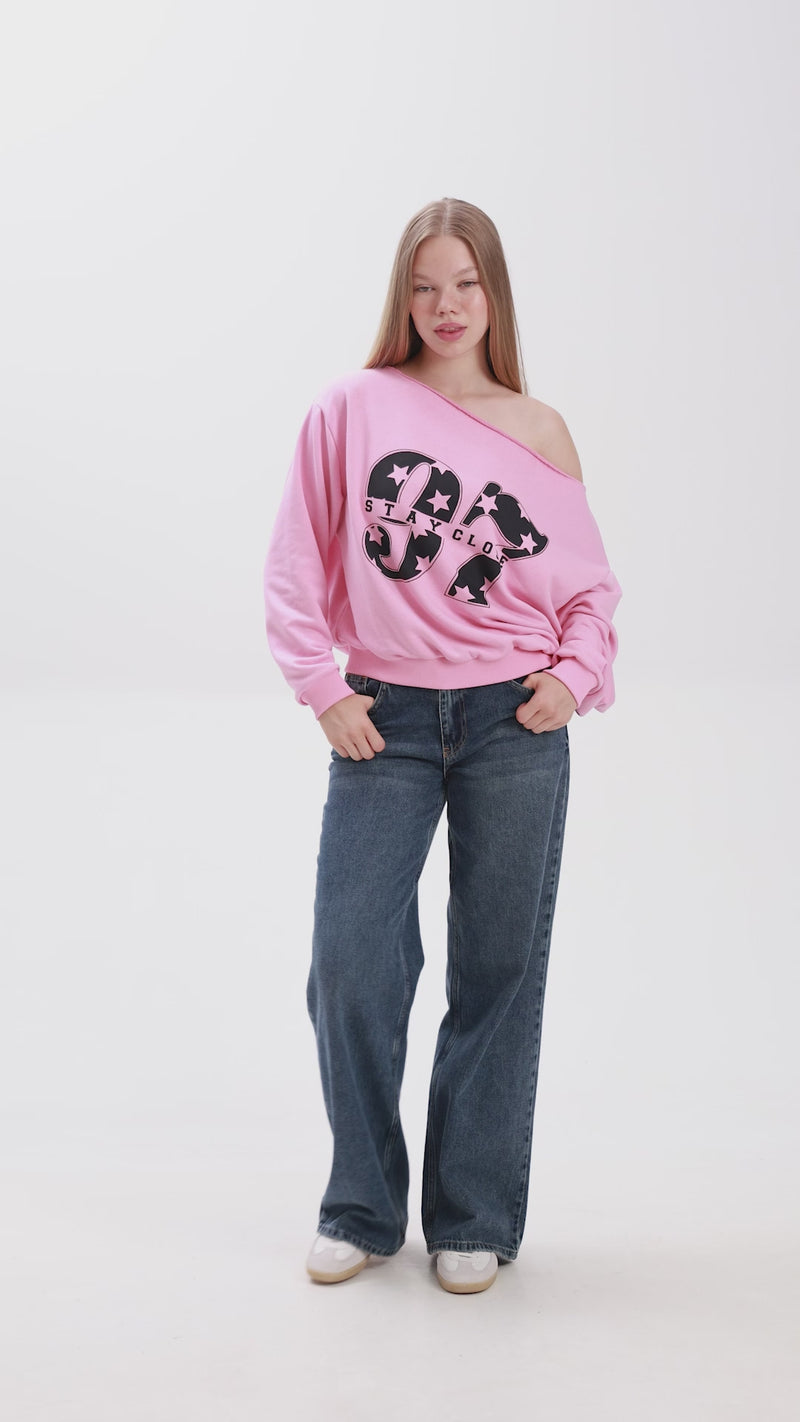 Model showcasing a pink off-shoulder sweatshirt with bold “Stay Close” print, paired with blue jeans and white sneakers.