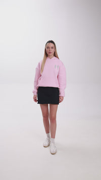 Studio video showcasing two hoodie options: navy and pastel pink. The models present the hoodies by moving, showing front and side angles, and highlighting details like the silver wing motif on the pink hoodie and the graphic logo on the navy hoodie. Styled casually with mini skirts and white sneakers for a modern streetwear look.